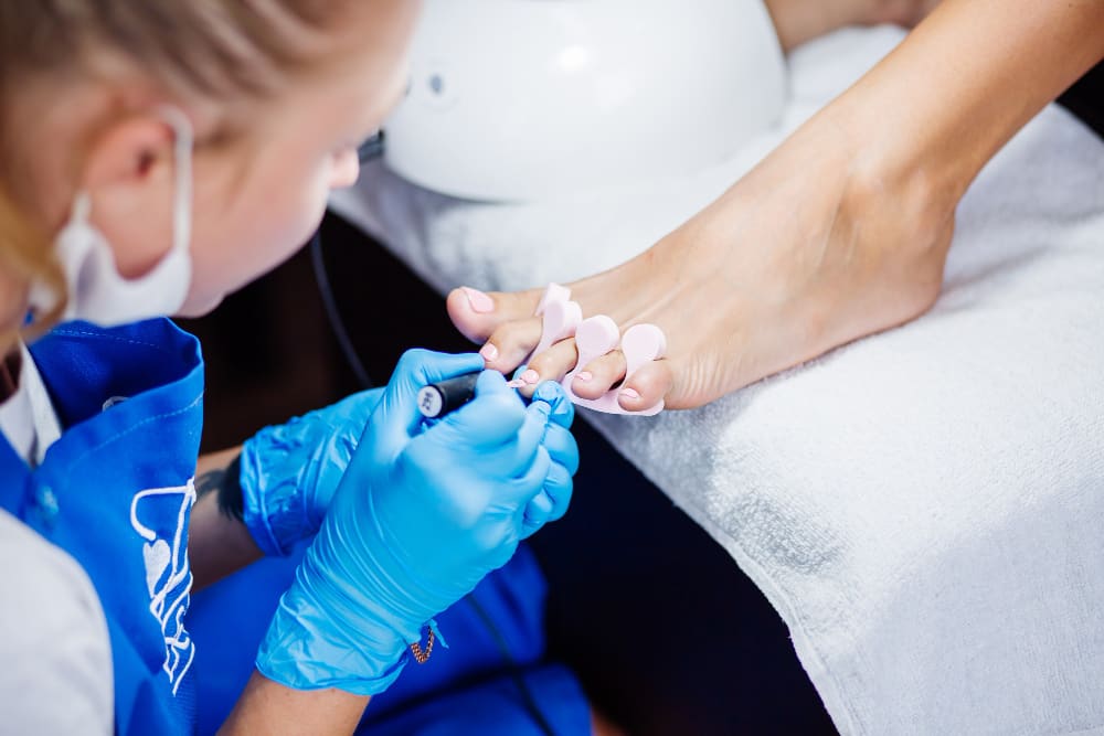Pedicurzystka w niebieskim fartuchu i rękawiczkach maluje paznokcie u stóp klientki, która ma założone różowe separatory międzypalcowe.