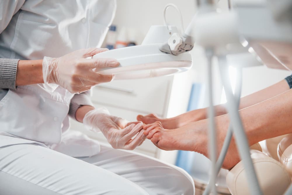 Specjalista podologii w białym fartuchu i rękawiczkach trzyma lampę z lupą powiększającą nad stopami klientki, aby dokładnie ocenić stan paznokci i skóry.